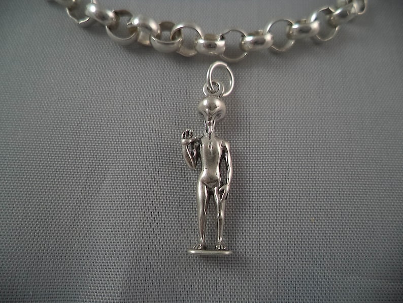 May include: A silver chain necklace with a silver alien charm. The alien is standing with one arm raised and is wearing a simple outfit.