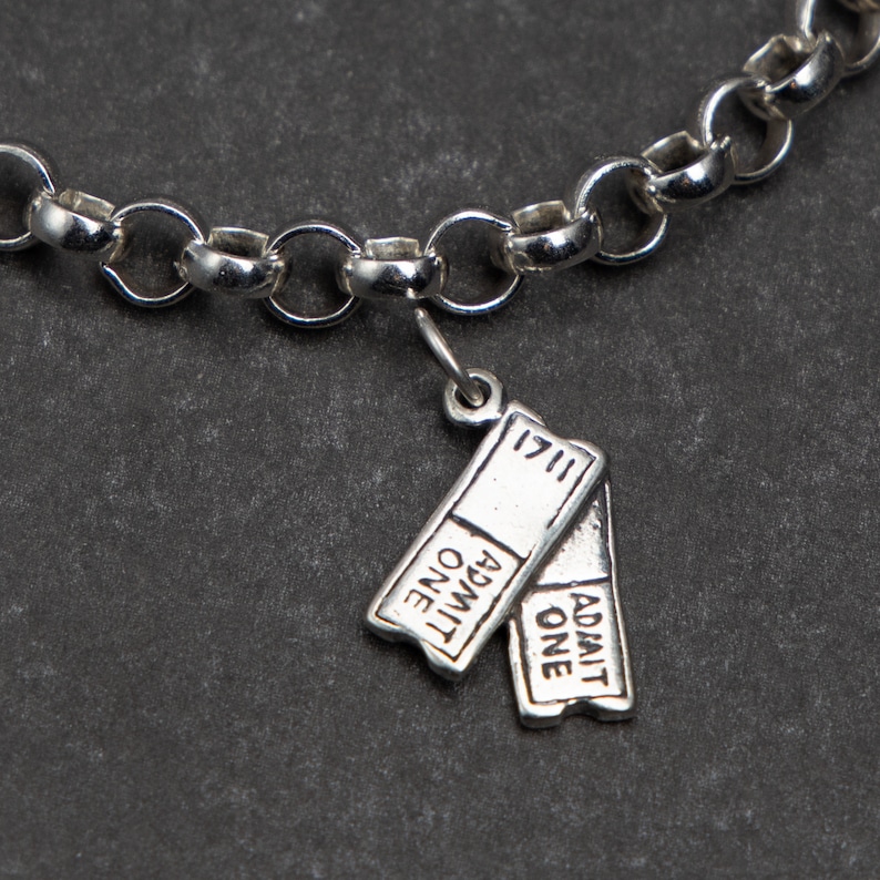 Puede incluir: Un brazalete de plata con un dije de billete de plata que dice "Admit One" en ambos lados. El dije est&aacute; unido a una cadena de plata.