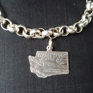 Op de afbeelding: Zilveren armband met een charme van de staat Washington met de tekst "WASHINGTON, SEATTLE, TACOMA, OLYMPIA, GRAND COULEE DAM".