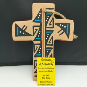 Puede incluir: Una cruz de terracota con un diseño nativo americano pintado en negro, blanco, azul y naranja. La cruz tiene un certificado de autenticidad que dice "Guaranteed Native American Made" y "Artist Tim Tosa Jemez Pueblo".