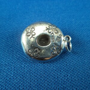 May include: Silver charm in the shape of a donut with four shamrock designs around the edge. The charm has a hole in the center and a loop for attaching to a bracelet or necklace.