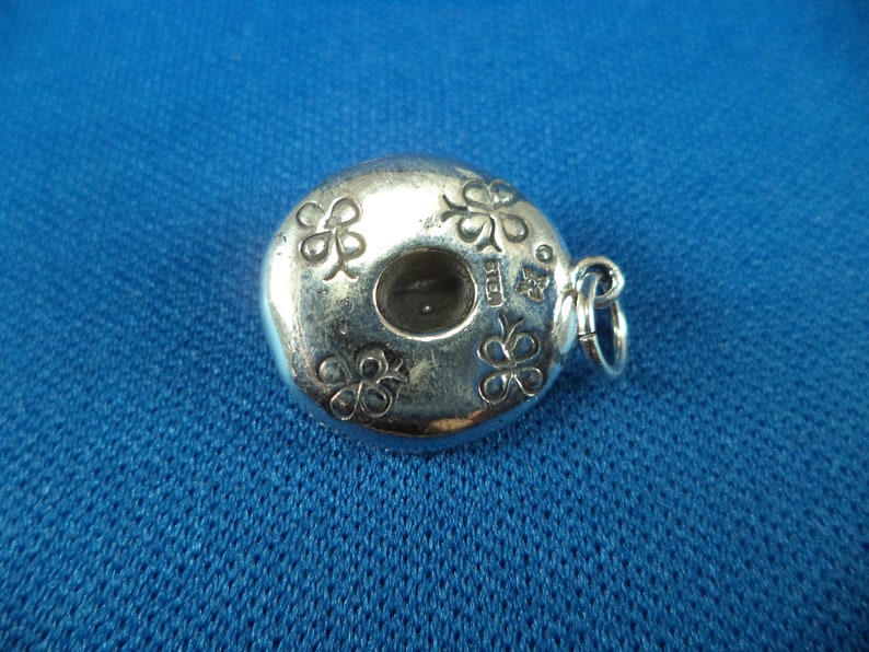 May include: Silver charm in the shape of a donut with four shamrock designs around the edge. The charm has a hole in the center and a loop for attaching to a bracelet or necklace.