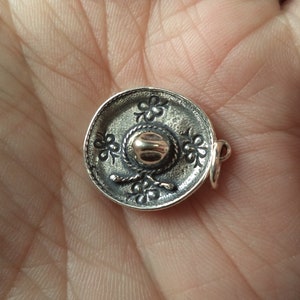 May include: A silver charm in the shape of a sombrero. The sombrero has a gold-colored band around the crown and is decorated with floral designs.
