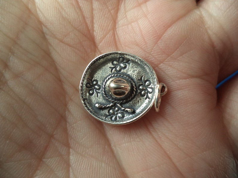 May include: A silver charm in the shape of a sombrero. The sombrero has a gold-colored band around the crown and is decorated with floral designs.
