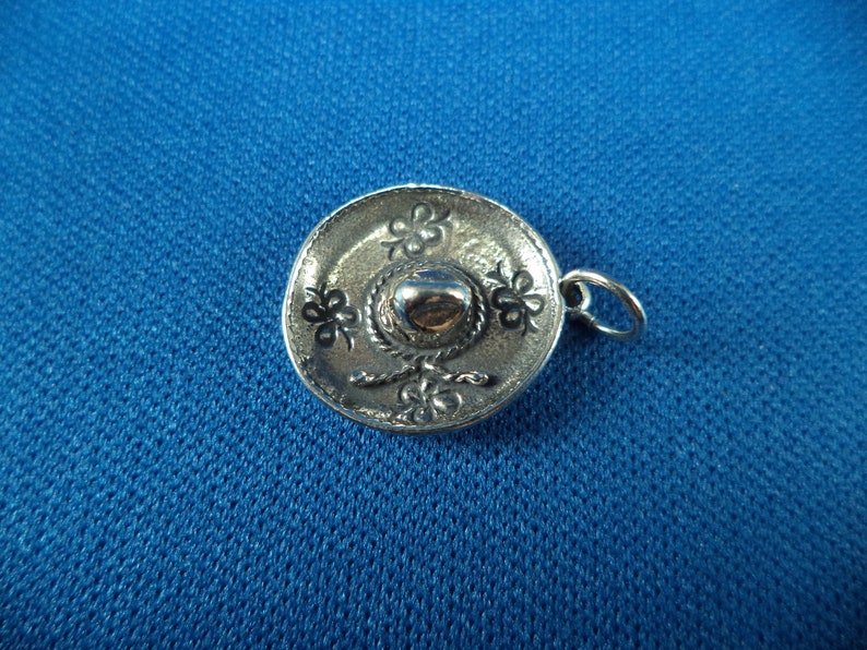 May include: A silver charm in the shape of a sombrero. The charm has a detailed design with a band and a flower pattern.