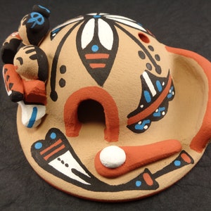 Può includere: Casa in ceramica di stile nativo americano dipinta a mano con una piccola apertura, decorata con motivi geometrici in nero, bianco, rosso e blu. La casa ha una piccola apertura ed è sormontata da tre piccole figure.