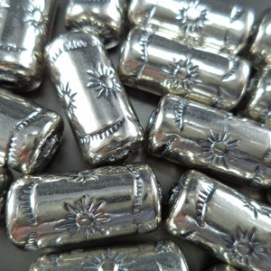 May include: Close-up of several silver-colored barrel beads. Each bead is cylindrical with a sunburst design and decorative edges. The beads are scattered, reflecting light and creating a textured appearance. Ideal for jewelry making.
