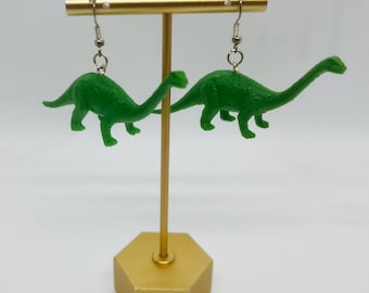 Könnte beinhalten: Ein Paar grüne Dinosaurier-Ohrringe, die an einem goldenen Ständer hängen. Die Dinosaurier haben lange Hälse und Schwänze.
