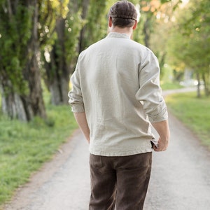 May include: A person wearing a light brown linen shirt and brown pants walks down a path lined with trees.