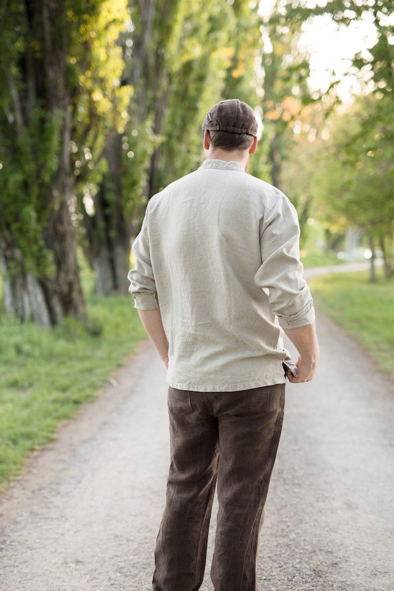 May include: A person wearing a light brown linen shirt and brown pants walks down a path lined with trees.