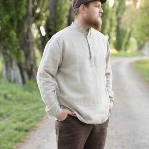 May include: A man wearing a brown cap and a light beige linen shirt with a button-up collar. He is wearing brown linen pants and has a beard. He is standing on a path in a forest.