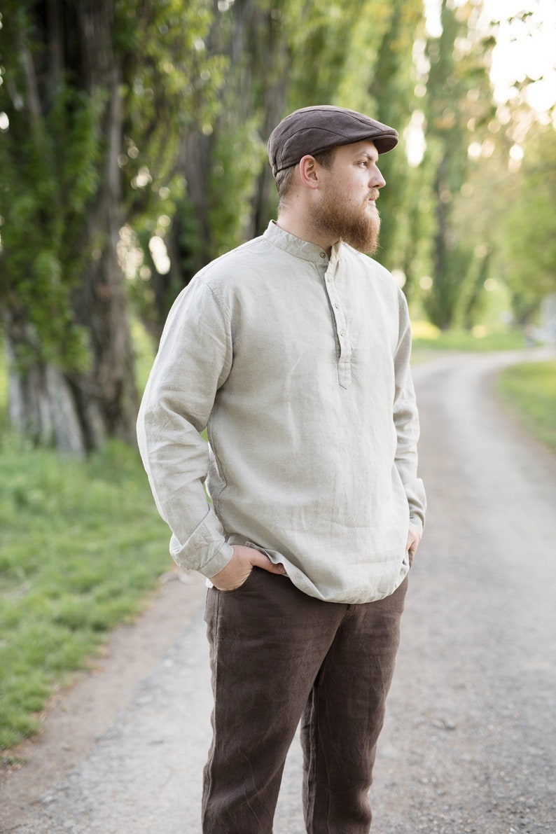 May include: A man wearing a brown cap and a light beige linen shirt with a button-up collar. He is wearing brown linen pants and has a beard. He is standing on a path in a forest.