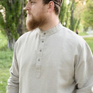 May include: A man wearing a brown newsboy cap and a light beige linen shirt with a button-up collar.