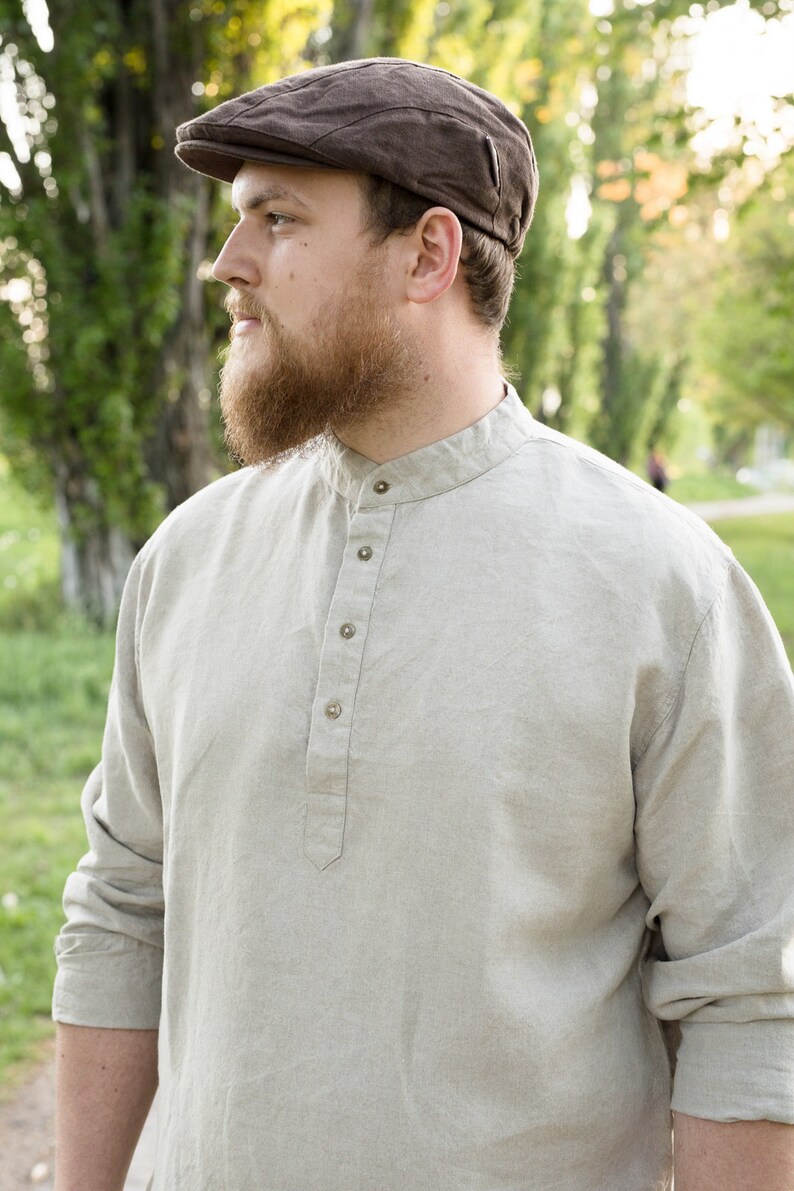 May include: A man wearing a brown newsboy cap and a light beige linen shirt with a button-up collar.