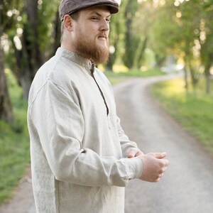 May include: A man wearing a brown cap and a light beige linen shirt with a collar. He is standing on a dirt path with green trees in the background.