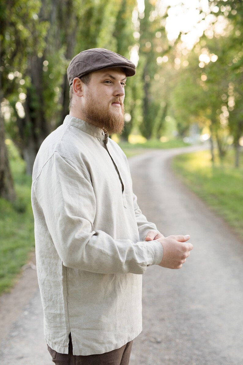 May include: A man wearing a brown cap and a light beige linen shirt with a collar. He is standing on a dirt path with green trees in the background.