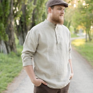 May include: A man wearing a brown cap and a light beige linen shirt with a button-up collar. He is standing on a path with trees on either side.