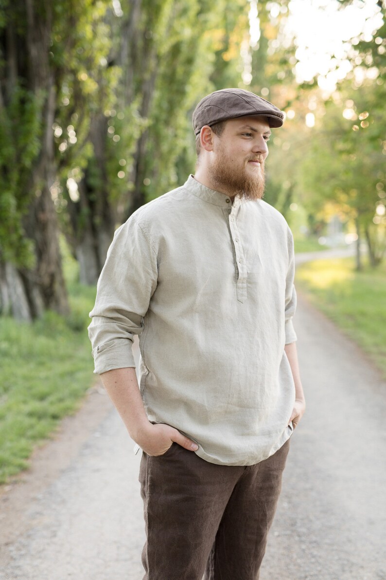 May include: A man wearing a brown cap and a light beige linen shirt with a button-up collar. He is standing on a path with trees on either side.