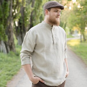 May include: A man wearing a light beige linen shirt with a mandarin collar and a brown newsboy cap. The shirt has a single button placket and long sleeves. He is standing in a wooded area with a path behind him.