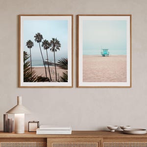 May include: Two framed wall art prints featuring coastal scenes. The left print shows palm trees, a sandy beach, and a pier extending into the ocean. The right print displays a solitary turquoise lifeguard tower on a vast, empty sandy beach.
