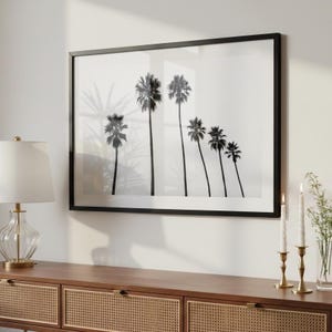 May include: A rectangular wall art piece featuring a black and white photograph of six palm trees of varying heights against a plain white background, enclosed in a thin black frame, displayed above a wooden console table with cane detailing.