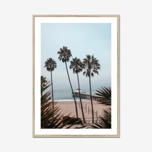 May include: A framed print featuring a beach scene with palm trees and a pier. The photograph has a muted color palette with a sandy beach, ocean, and a cloudy sky. The palm trees are silhouetted against the sky.