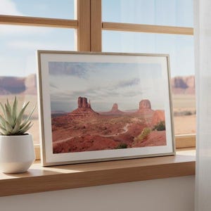 May include: Framed landscape print featuring Monument Valley's iconic red rock formations under a cloudy sky. The print is displayed in a light-colored wooden frame, resting on a wooden windowsill. A potted succulent plant is visible on the left.