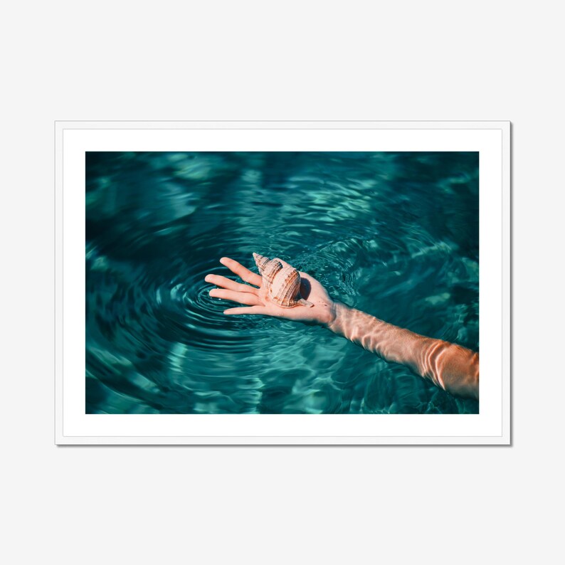 Coastal decor wall art showcasing a seashell and hand submerged in clear blue green ocean water. This giclée photography print highlights texture, light, and movement, perfect for serene interiors and summer inspired home styling.