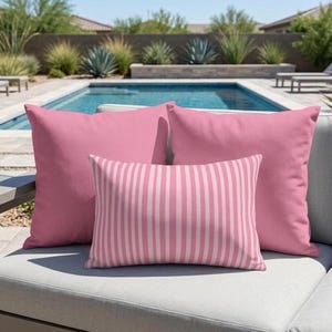 May include: Two square solid pink throw pillows and one rectangular throw pillow featuring a vertical light pink and white striped pattern. The pillows are arranged together on a light gray outdoor cushion surface.