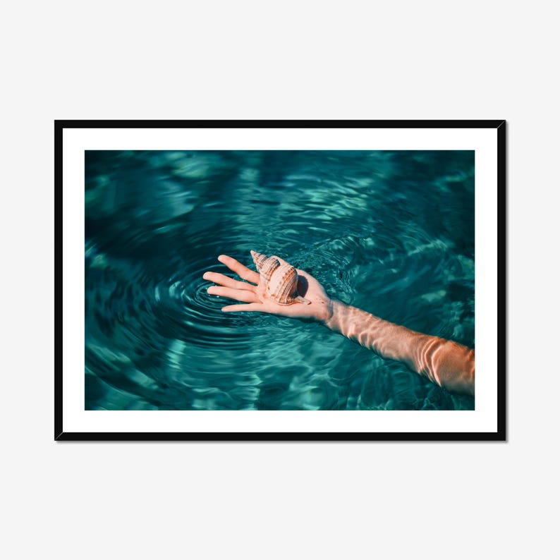 Teal beach photography print featuring an outstretched hand holding a seashell in calm ocean water. This framed coastal wall art captures a peaceful seaside moment and works beautifully in beach house decor, modern coastal homes, or ocean themed spaces.
