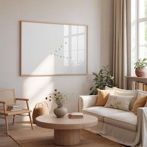 May include: A large rectangular wall art piece in a light wood frame featuring a minimalist white background with a small black graphic of a flock of birds flying in a V-formation. The artwork is displayed on a plain wall above a living room.