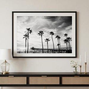 May include: A black and white framed landscape photograph depicting a long pier extending into the ocean, lined with tall palm trees under a cloudy sky. The artwork is displayed in a thin black frame, showcasing a coastal scene with silhouettes.