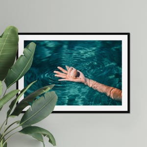 Teal ocean photography framed as fine art wall decor, featuring a hand holding a seashell in shallow water. This beach themed print suits coastal living rooms, bedrooms, entryways, or seaside vacation homes.