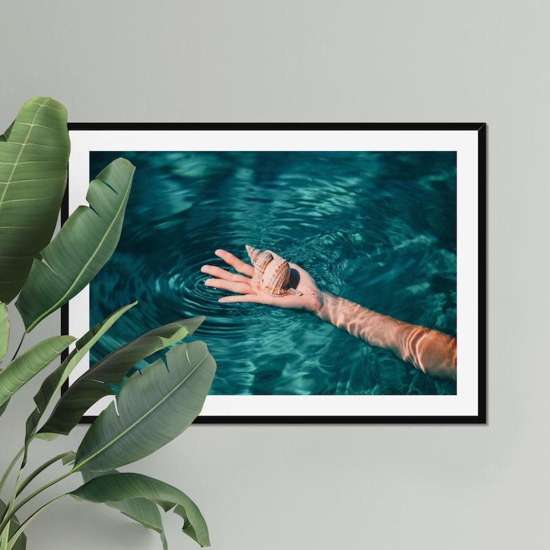 Teal ocean photography framed as fine art wall decor, featuring a hand holding a seashell in shallow water. This beach themed print suits coastal living rooms, bedrooms, entryways, or seaside vacation homes.