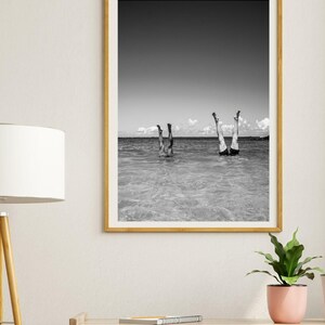 May include: Black and white photograph of two people standing on their hands in the ocean. The photo is framed in a light wood frame.