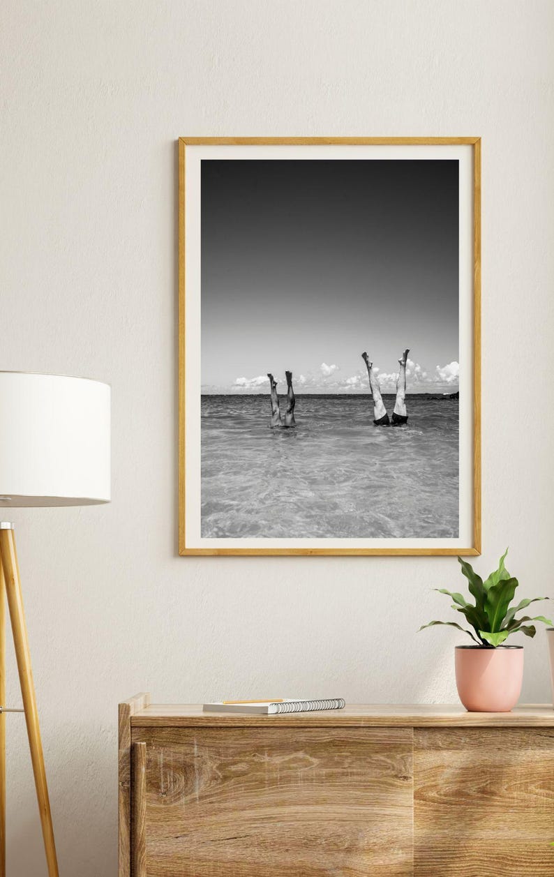 May include: Black and white photograph of two people standing on their hands in the ocean. The photo is framed in a light wood frame.