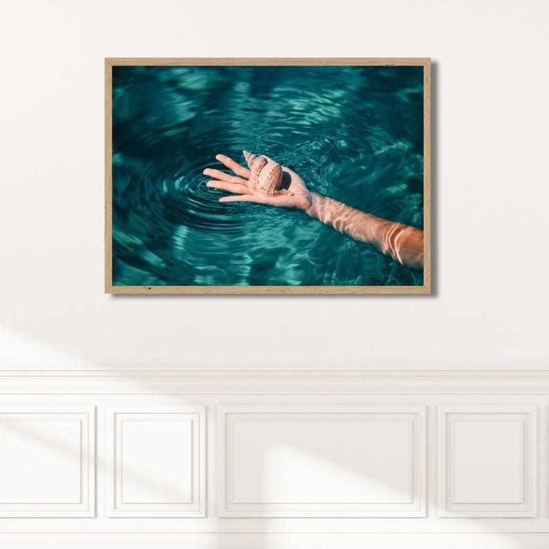 Seaside wall art print with a minimalist composition of a seashell resting in a hand surrounded by calm ocean ripples. This framed coastal artwork brings a peaceful, airy mood to modern beach house decor. Fine art coastal photography print featuring a seashell and gentle water movement in vibrant teal tones. This framed ocean wall art is ideal for summer decor, coastal interiors, and relaxed seaside inspired spaces.