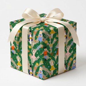 May include: A square gift box wrapped in dark green paper featuring a repeating pattern of nutcrackers, ballerinas, and pine branches, tied with a wide, cream-coloured satin ribbon bow on top. The design includes small stars and bows.