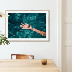 Framed beach wall art featuring a sea shell held above rippling teal water. This coastal photography print adds an elegant seaside feel to home decor, blending fine art style with a relaxed ocean inspired aesthetic.