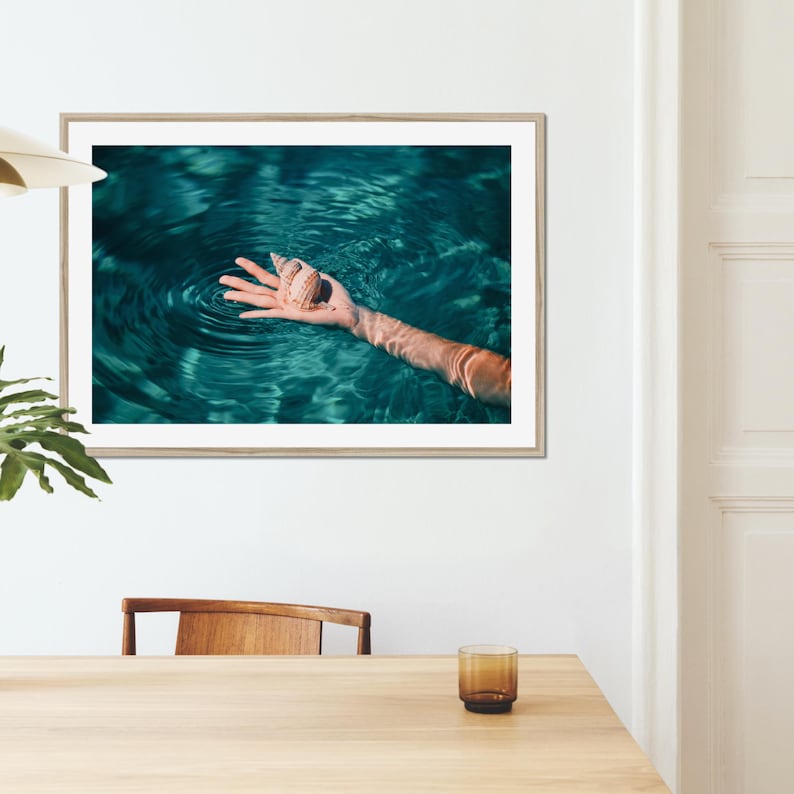 Framed beach wall art featuring a sea shell held above rippling teal water. This coastal photography print adds an elegant seaside feel to home decor, blending fine art style with a relaxed ocean inspired aesthetic.