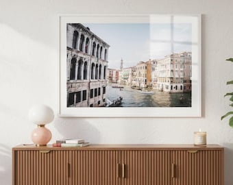 May include: A framed landscape photograph of a Venetian canal scene featuring historic multi-story buildings with arched windows, small boats on the water, and a distant bell tower under a clear sky, displayed in a white frame on a wall.