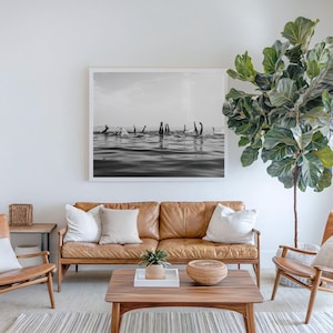 May include: A black and white framed photograph depicting multiple pairs of legs sticking out of the water, mounted on a white wall above a tan leather sofa, two wooden armchairs, a wooden coffee table, and a large potted fiddle leaf fig tree.