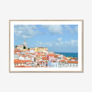 May include: A framed print of a cityscape featuring buildings with red tile roofs, set against a blue sky with fluffy white clouds. The buildings are painted in various colors, including yellow, pink, and white, with a domed structure in the background.