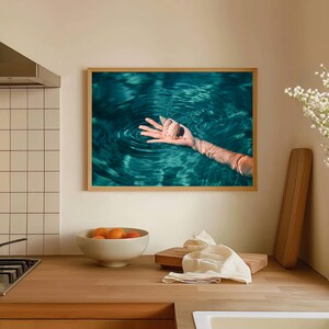 Teal beach photography print featuring an outstretched hand holding a seashell in calm ocean water. This framed coastal wall art captures a peaceful seaside moment and works beautifully in beach house decor, modern coastal homes, or ocean themed spaces.