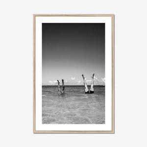 A black and white photograph of two people standing on their hands in the water. The water is shallow and the sky is clear. The people are wearing swimsuits and are facing away from the camera. Black and white beach scene photo framed in natural wood, vertical wall art adds vintage coastal charm to a beach house.