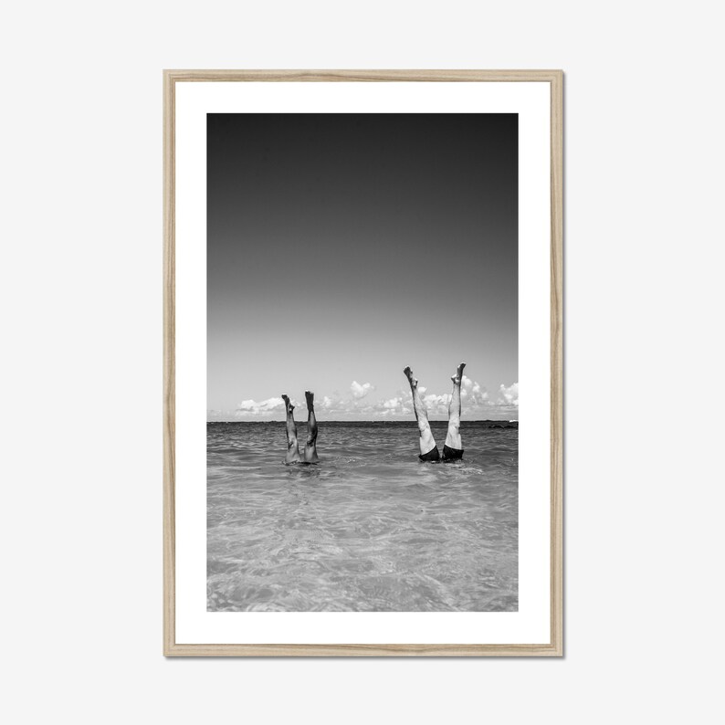 A black and white photograph of two people standing on their hands in the water. The water is shallow and the sky is clear. The people are wearing swimsuits and are facing away from the camera. Black and white beach scene photo framed in natural wood, vertical wall art adds vintage coastal charm to a beach house.
