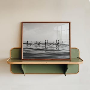 May include: A black and white photograph in a dark wood frame resting on a green and light wood wall-mounted shelf. The image depicts multiple pairs of legs extending vertically out of the water, capturing a scene of swimmers diving or playing.