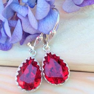 May include: A pair of silver teardrop earrings with red crystal stones. The earrings have a delicate, ornate design.