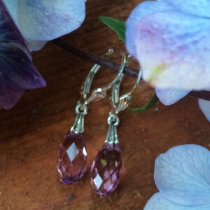 May include: A pair of silver hoop earrings with teardrop-shaped amethyst crystals hanging from them. The earrings are photographed against a background of purple and blue flowers.