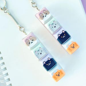 May include: Two keychains with silver hardware and spring coils. Each keychain features a stack of five rectangular keycaps with cat faces in various colors. The keychains rest on a white spiral notebook against a light purple background.